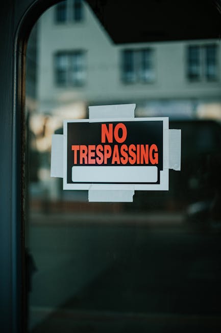 Close-up of a printed 'No Trespassing' sign affixed to a glass door or window, with several pieces of beige or white tape securing it around the edges. The sign features bold red and black text on a black background, indicating restricted access to the property. In the blurred background, parts of a residential building with multiple windows and a light-colored facade are visible, suggesting the setting is in an urban or suburban area. The presence of the sign and tape indicates a security measure or restricted entry, which may be relevant during house removal or relocation services to ensure safety and property protection. This scene demonstrates the importance of clear boundary marking when conducting home or furniture transport, a common aspect of professional removals undertaken by services like Man with Van South Hornchurch.