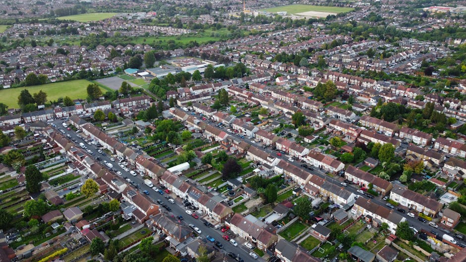 Aerial view of a residential neighbourhood in South Hornchurch showing rows of terraced houses with small front gardens, some with parked cars along the street. Visible are various cardboard boxes, plastic wrapping, and furniture coverings indicating a furniture transport or home relocation process. The loading area includes a white van belonging to Man with Van South Hornchurch parked near the houses, with some boxes and packing materials visible inside or nearby. The houses are situated in a suburban setting with green trees and open spaces in the background, and the scene appears to be during daytime with natural light illuminating the area, capturing the typical layout and logistics involved in house removals and packing during a move.