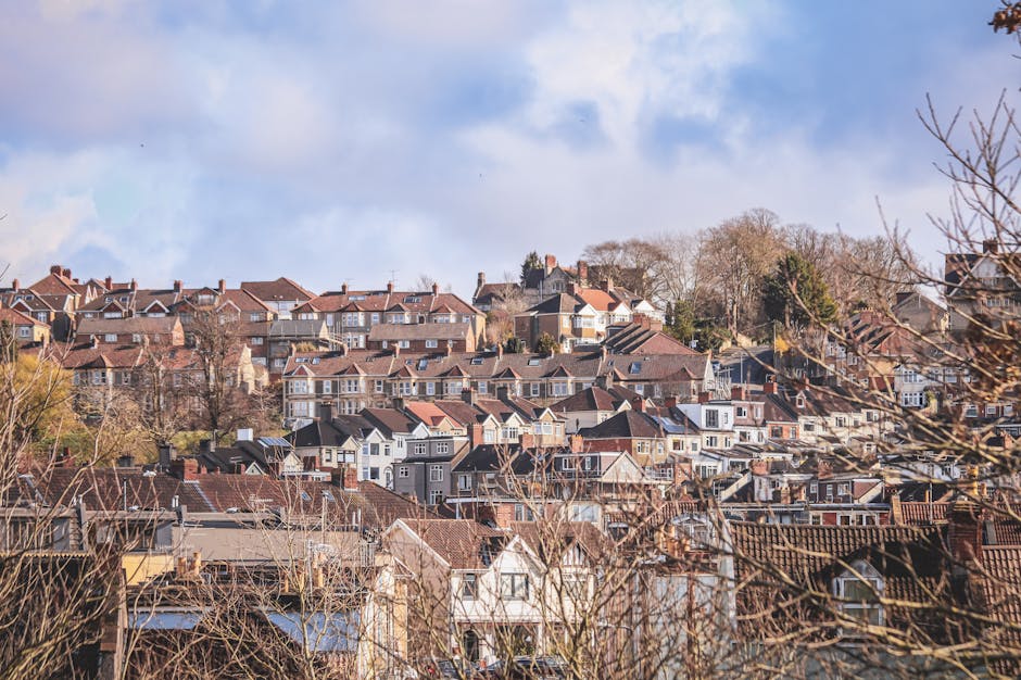 A panoramic view of a residential area in South Hornchurch showing multiple closely packed houses with varying architectural styles, predominantly two to three stories, with tiled roofs in shades of red, brown, and gray. Many homes feature white or pale-colored facades, large windows, and some have chimneys. The area is situated on a hilly terrain, with houses ascending towards the background, surrounded by leafless trees and shrubbery indicating late autumn or winter season. In the foreground, there are some bare branches and rooftops, while the sky is mostly cloudy with patches of blue. This scene captures a typical suburban neighborhood where property owners and movers like Man with Van South Hornchurch often assist with furniture transport and home relocation processes through loading and packing activities, sometimes involving visible cardboard boxes and protective covers on furniture, preparing for an efficient house removal or moving service.