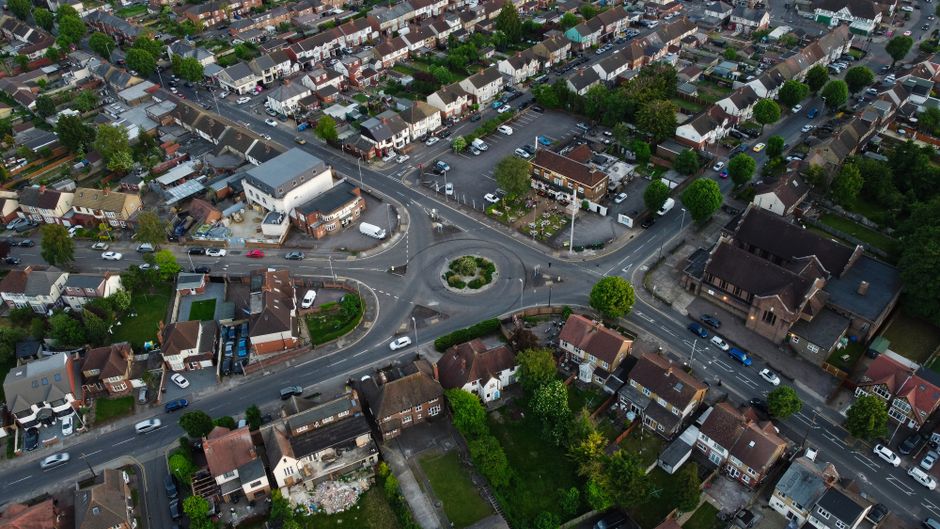 Aerial view of a residential neighbourhood in South Hornchurch showing rows of terraced houses with small front gardens, some with parked cars along the street. Visible are various cardboard boxes, plastic wrapping, and furniture coverings indicating a furniture transport or home relocation process. The loading area includes a white van belonging to Man with Van South Hornchurch parked near the houses, with some boxes and packing materials visible inside or nearby. The houses are situated in a suburban setting with green trees and open spaces in the background, and the scene appears to be during daytime with natural light illuminating the area, capturing the typical layout and logistics involved in house removals and packing during a move.