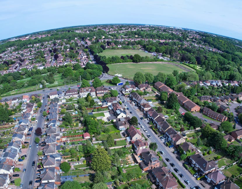 Aerial view of a suburban neighbourhood showing rows of terraced and semi-detached houses with front gardens, tree-lined streets, and parked cars. The scene includes a school playing field with green 