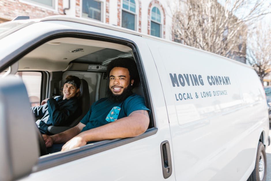 Inside the driver and passenger seats of a white moving van operated by Man with Van South Hornchurch, a smiling African American male driver with a beard and casual clothing is seated in the driver's seat, while a young woman with light skin, dark hair tied back with a headband, wearing a black jacket, is seated in the passenger seat. The van's interior shows part of the dashboard and ceiling, with the door open revealing the sidewalk and a residential street in the background, where trees and multi-story buildings are visible. The van displays the company's name and services, including local and long-distance removals, printed on the side. The scene depicts the loading process of home relocation, with the vehicle positioned outside a property, emphasizing furniture transport, packing, and moving logistics. The natural daylight enhances clarity of the objects inside and outside the van.