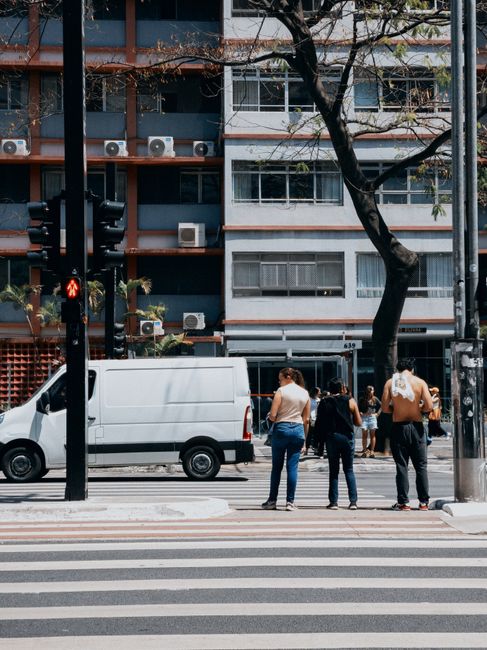 A group of three people standing on a pedestrian crossing at a street corner in front of a white panel van parked adjacent to a multi-storey residential building. The van is positioned with its side door closed, and is situated near a curb with the vehicle’s rear slightly visible. The individuals are dressed casually; one woman in a sleeveless top and jeans, another person with a dark top and glasses, and a third person wearing a white shirt with a backpack. The scene is illuminated by daylight, with a traffic light showing a red pedestrian signal, and a tree with bare branches partially obscuring the building. The background features the apartment block with multiple windows, air conditioning units, and a mix of brick and painted facades, consistent with hotel or apartment building architecture. This setting reflects the typical logistics involved in home relocation, furniture transport, or packing and moving activities managed by [COMPANY_NAME].