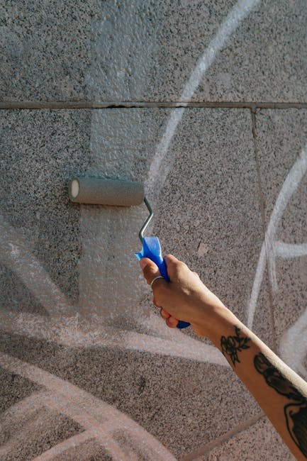 A person's hand, featuring a tattoo on the wrist, holding a paint roller with a blue handle as it applies light pink or beige paint onto a grey brick wall. The roller is positioned near the top left of the image, and the wall's surface displays visible mortar lines and textured brick patterns. The scene is indoors, illuminated by natural or artificial light, and is part of a home refurbishment or decorating process. The action of painting suggests ongoing interior work in a property scheduled for house removals or a furniture transport setup, which is managed by Man with Van South Hornchurch. The image captures the preparation or updating phase prior to packing or moving activities, emphasizing the importance of property readiness in professional relocation services.