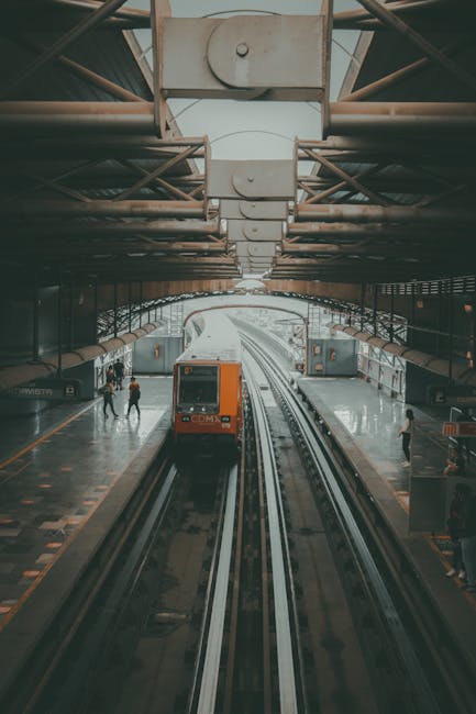 Image showing a modern indoor train station with an orange train approaching on the tracks, under a large, arched metal roof structure supported by beams. The platform is populated with a few passenge